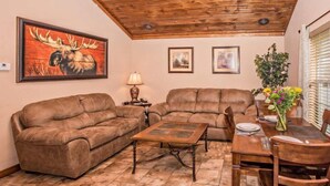 A cozy living room with two brown leather sofas, a wooden coffee table, and a dining table with chairs. There is a large painting of a moose on the wall, and a window with blinds to the right.