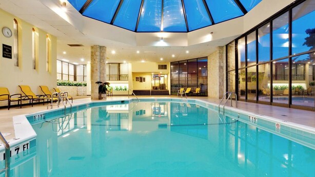An indoor swimming pool with a glass ceiling, yellow lounge chairs, and a stone pillar. - Reading Vitality Hotel (Wyomissing)