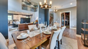 A spacious dining area with a wooden table set for six, surrounded by modern chairs. A chandelier hangs above, and the room features a high ceiling and a staircase leading to an upper level. - Spacious Lake Charles Craftsman - Pet Friendly! (Lake Charles)