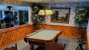 Une table de billard dans une pièce aux murs lambrissés, avec un téléviseur et un tableau représentant un paysage, encadré.
