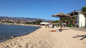 A beach with a thatched umbrella, a building with a modern design, and a view of mountains in the background. - Apartment Oleander - One Bedroom Apt with Terrace (Novalja)