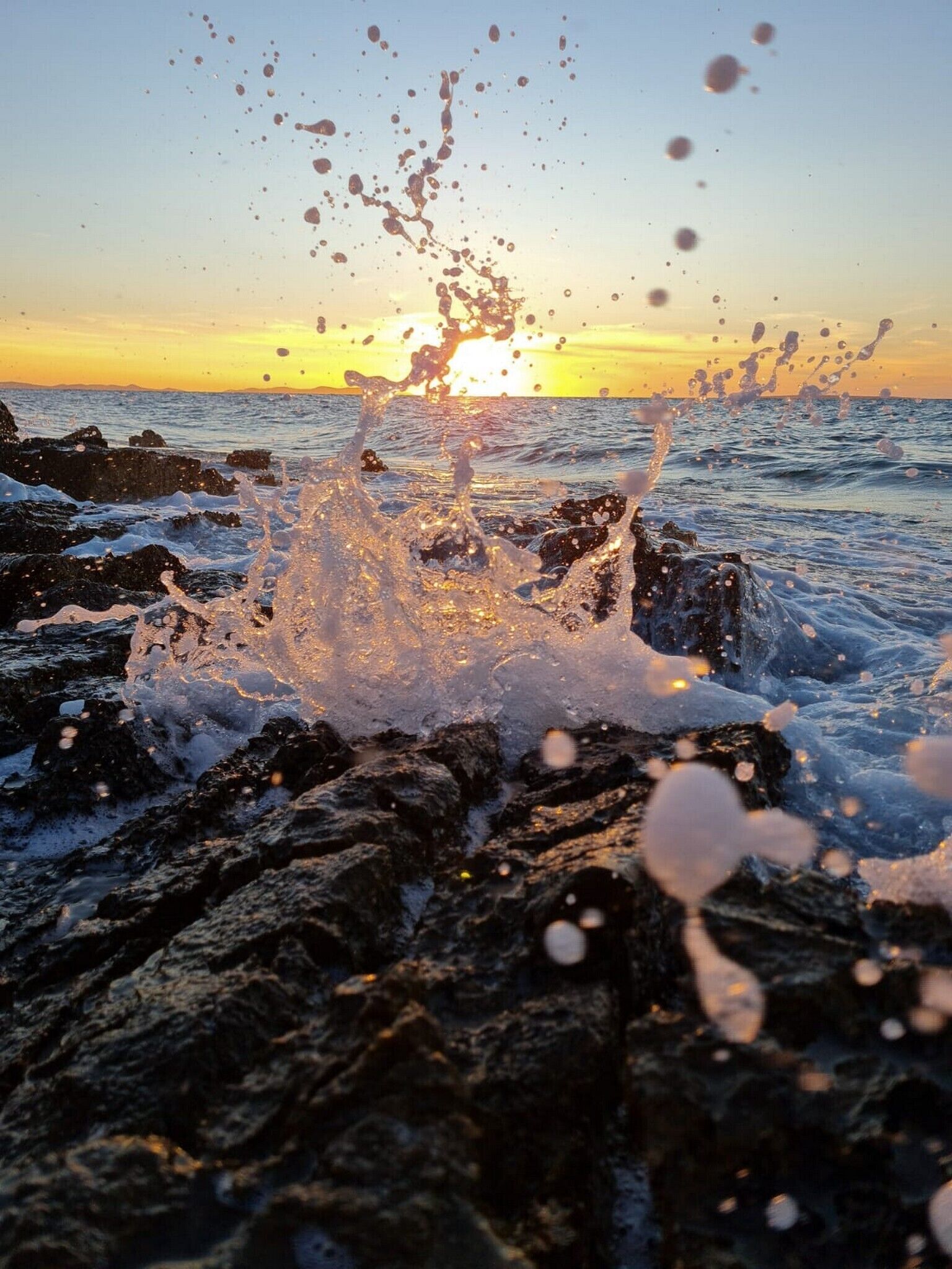 Auringonlasku kivikkoisella rannikolla, jossa aallot iskeytyvät kallioihin.