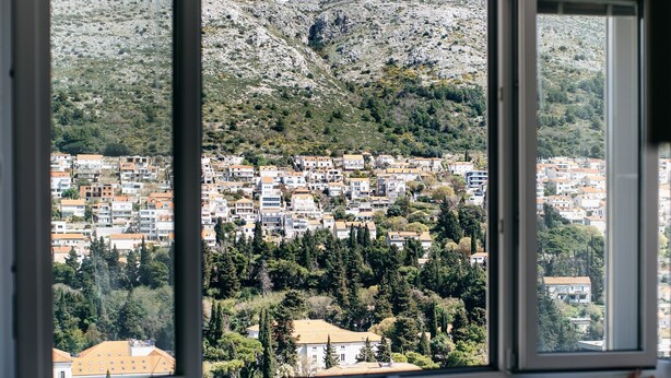 A dining area with a table and chairs, a window with a view of a town and hills. - Apartment Wanda - Two Bedroom Apartment with City View (Dubrovnik)