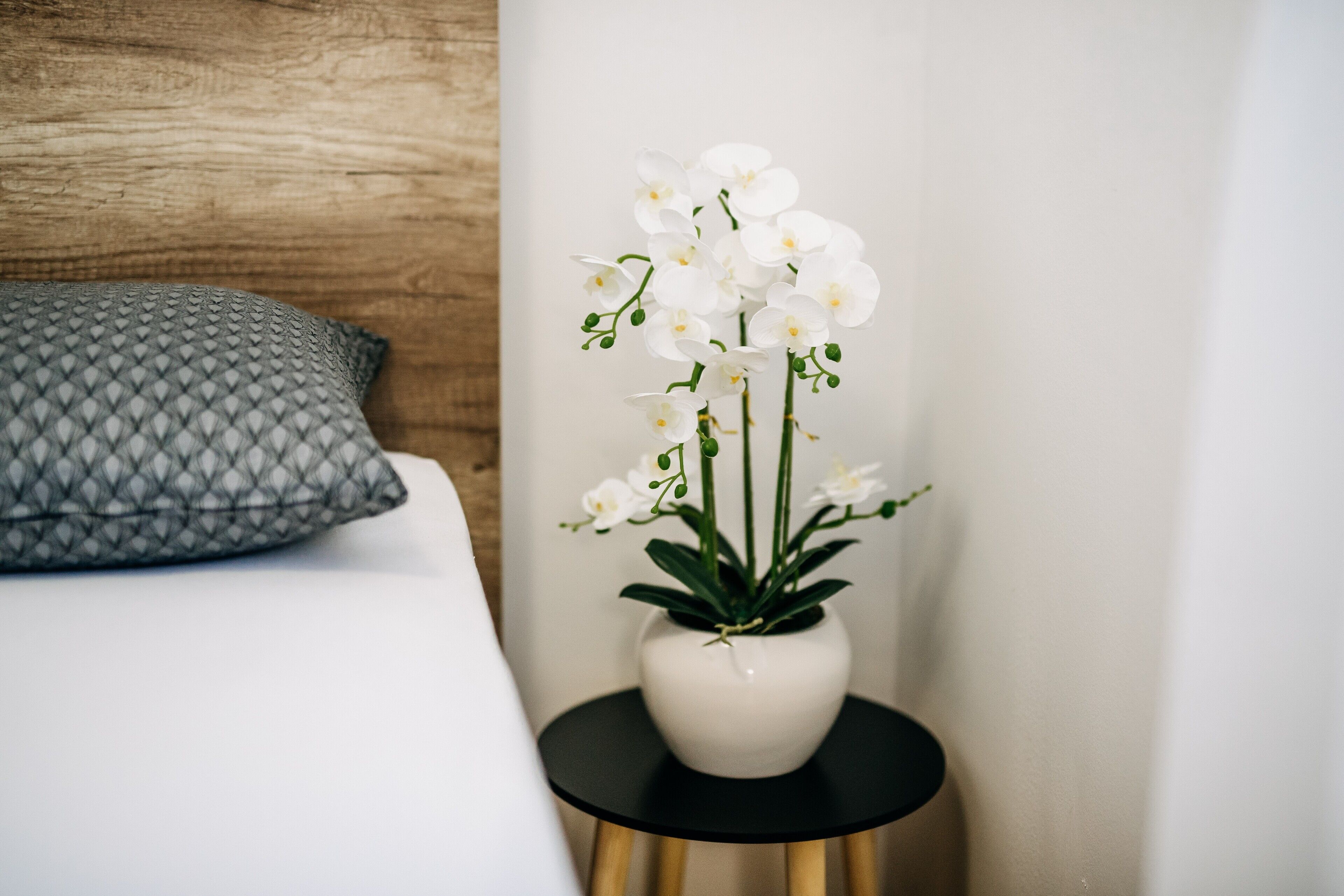 Una cama con un cojín estampado, un jarrón blanco con flores blancas sobre una mesita negra, y un cabecero de madera.
