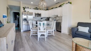 Eine moderne Küche mit weißen Schränken, einem Edelstahlkühlschrank, einem Glastisch mit weißen Stühlen und einem grauen Sofa im Wohnbereich.