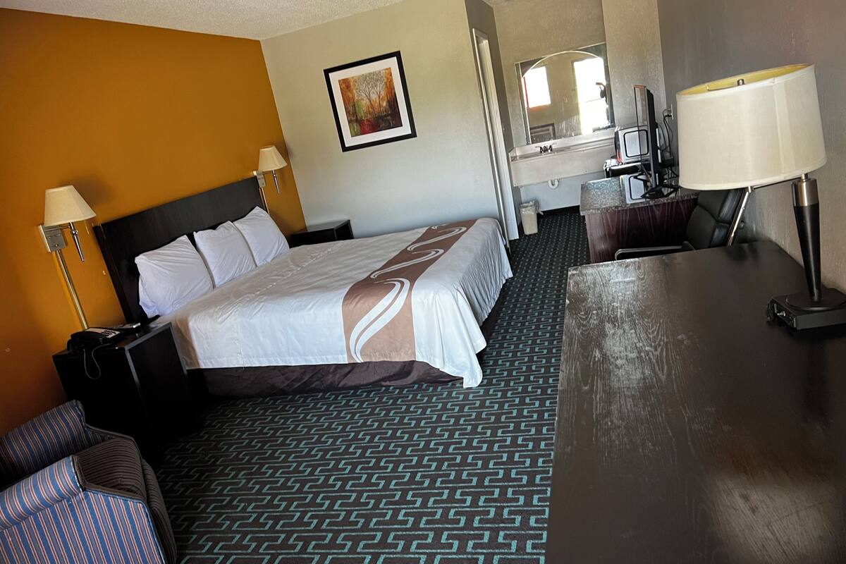 Quarto de hotel com uma cama grande, uma escrivaninha, uma cadeira, um abajur, um quadro na parede e um tapete estampado.