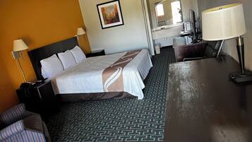A hotel room with a large bed, a desk, a chair, a lamp, a painting on the wall, and a patterned carpet.