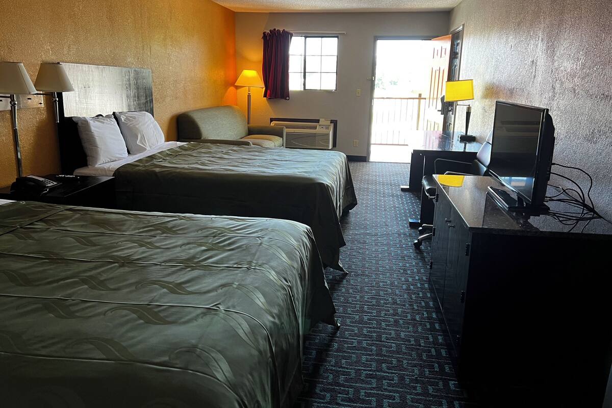 Quarto de hotel com duas camas, uma escrivaninha com computador, uma cadeira, um abajur e uma janela com cortinas.