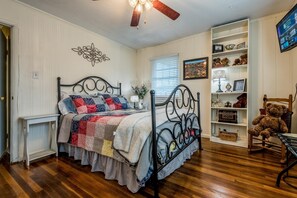 A bedroom with a bed, a ceiling fan, a bedside table, a nightstand, a window with blinds, a shelf with decorative items, and a teddy bear.