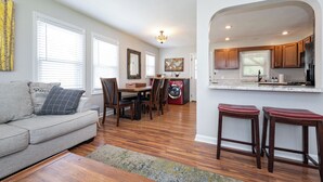 A living room with a sofa, dining table, and chairs, a kitchen with wooden cabinets and a refrigerator, and a laundry area with a washing machine. - OPEN 4BR/2BA • Sleeps 8 • Near Hwy • Covered Deck • Close to Downtown & Expo Ctr (Louisville)
