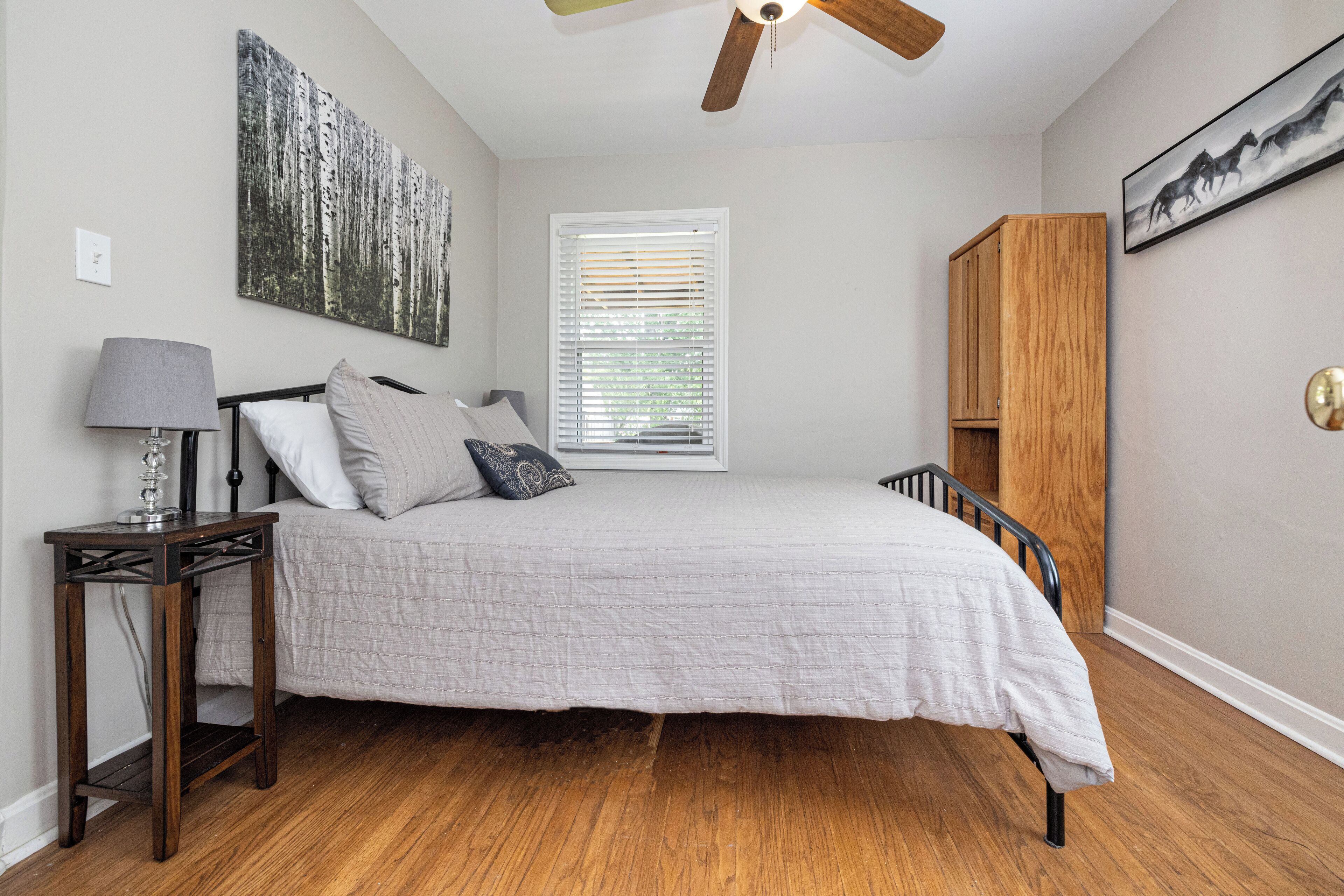 Une chambre à coucher comprenant un lit, une table de chevet, une armoire en bois, un ventilateur de plafond et une fenêtre avec des stores.