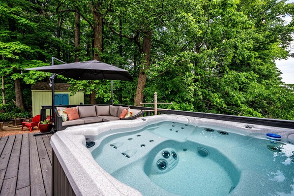 Un jacuzzi rodeado por una terraza de madera, con un sofá y sillas bajo un toldo, con un fondo de árboles frondosos.