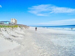 Une plage avec une personne qui se promène, une jetée et des bâtiments en arrière-plan.