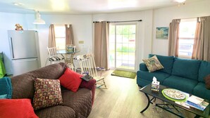 A living room with a brown sofa, teal sofa, and a glass coffee table. There is a refrigerator, a dining table with chairs, and a sliding glass door leading to an outdoor area. - ❤️Amazing Round House, Ground Floor Unit, Easy Walk To The Beach, Self Check-In (Carolina Beach)