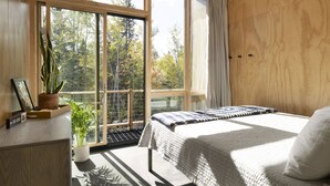 A bedroom with a large window, a bed, a bedside table, and a potted plant.