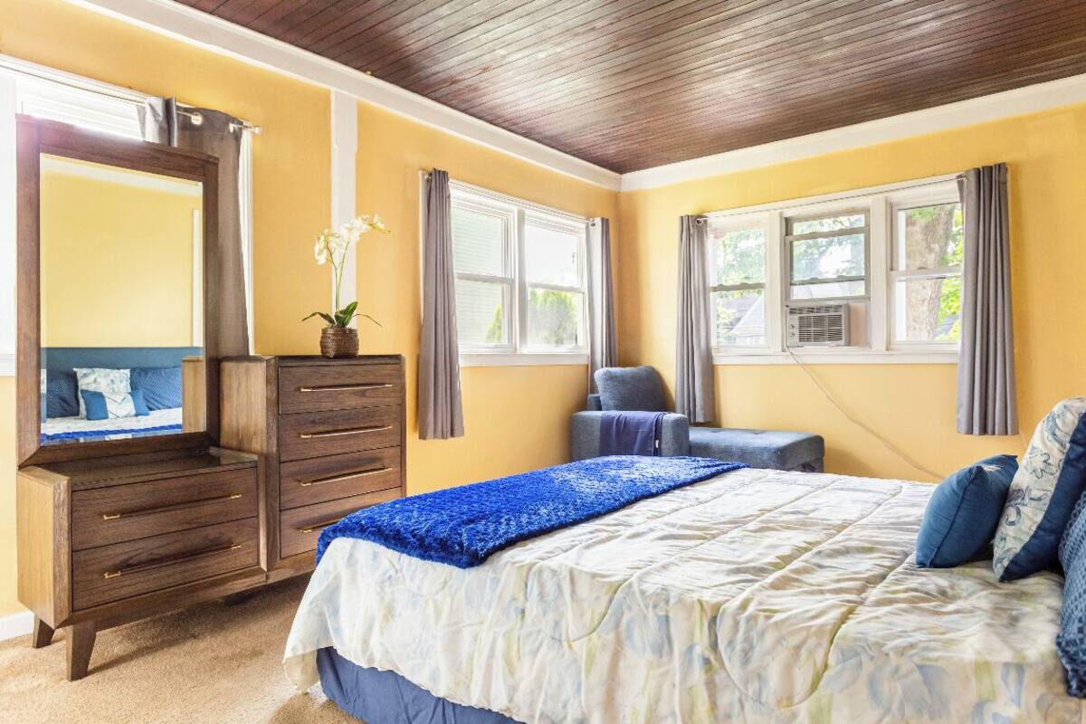 A bedroom with a wooden dresser, a bed with blue bedding, and two windows with curtains.