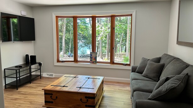 A modern living room with a large window, a grey sofa, a wooden coffee table, and a TV mounted on the wall.