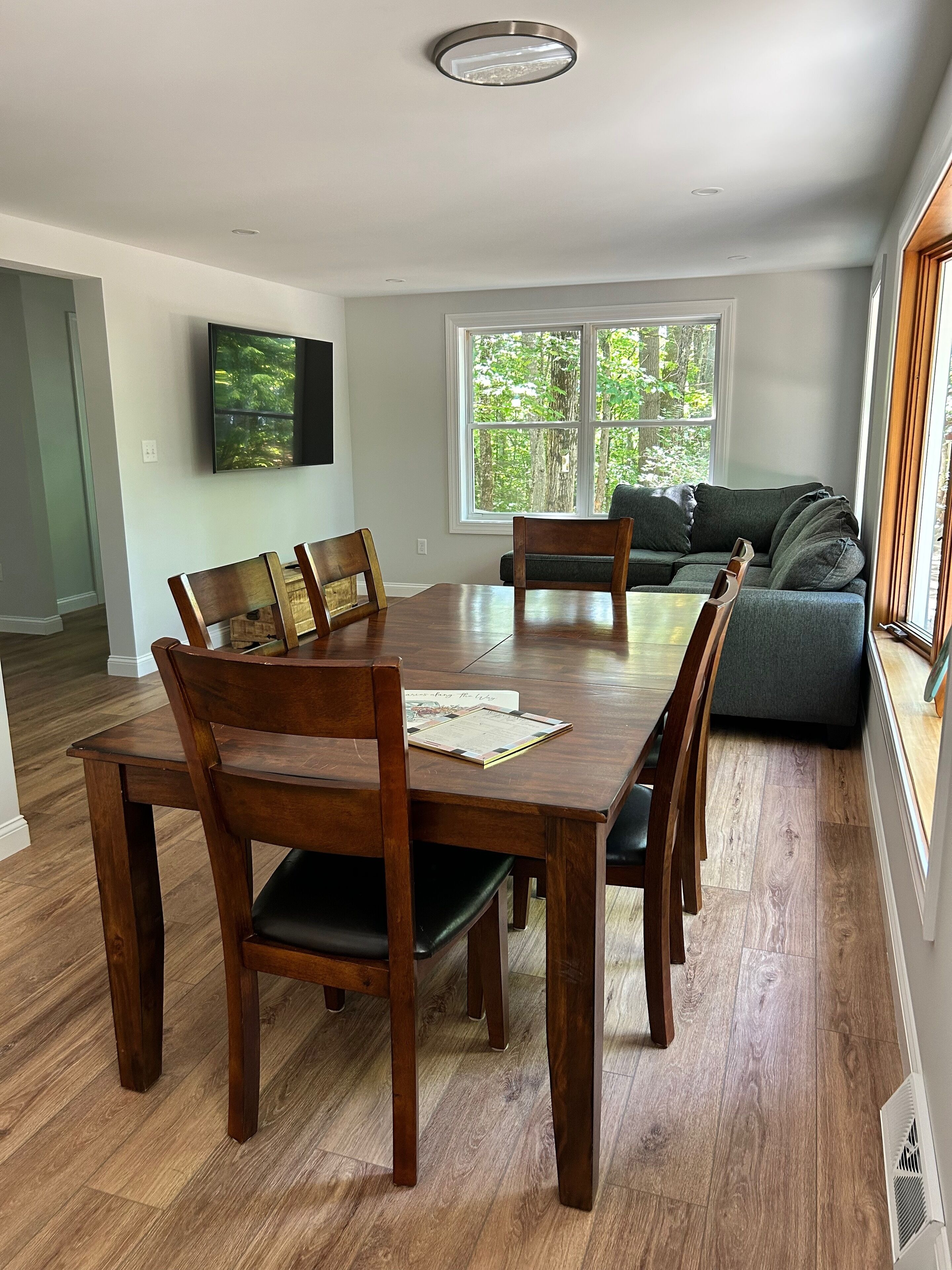 A dining area with a wooden table and chairs, a sofa, and a large window with a view of trees.