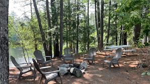 A campsite with Adirondack chairs arranged around a fire pit, overlooking a lake.