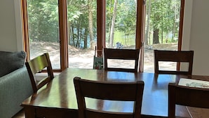 A dining area with a wooden table and chairs, large windows with a forest view, and a grey sofa in the background.