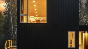 A modern, compact building with a large window revealing a room with a bed and seating area. - Scandinavian Chalet and sauna in Lutsen - Grey (Lutsen)