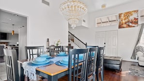 Un'area pranzo con tavolo di legno apparecchiato per sei persone, circondato da sedie nere. Un lampadario pende dal soffitto, e al muro è appeso un quadro.
