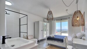 Une salle de bain moderne avec un lavabo blanc, une douche et une grande fenêtre donnant sur la plage.