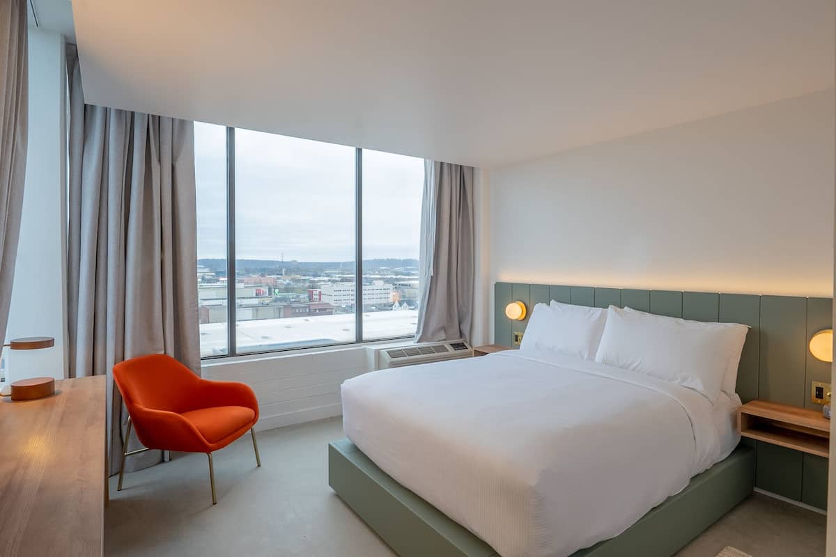 Une chambre d’hôtel avec une grande fenêtre, un lit, une chaise et un bureau.