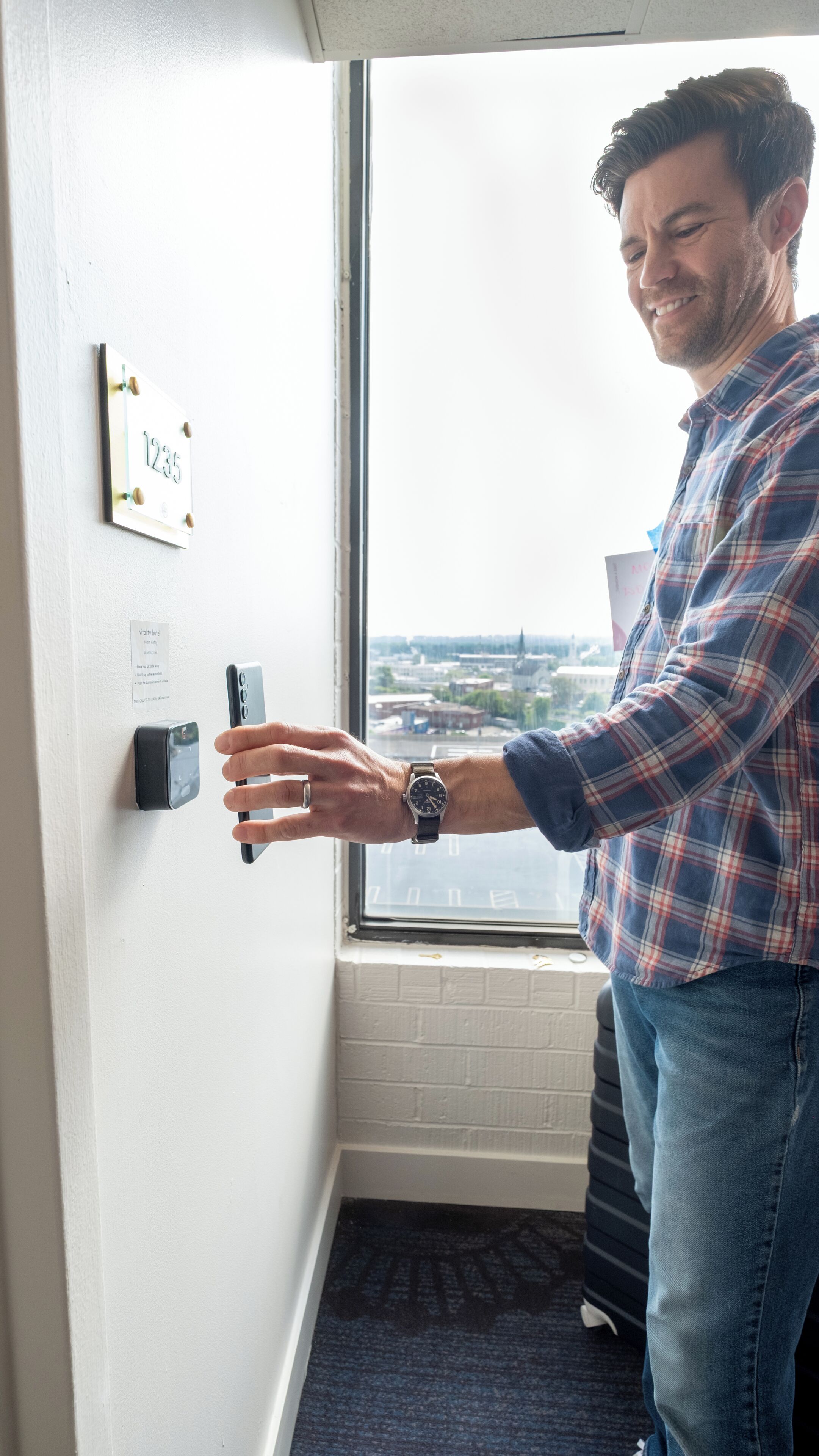 A person unlocking a door with a key card, a room number sign, and a window with a city view.