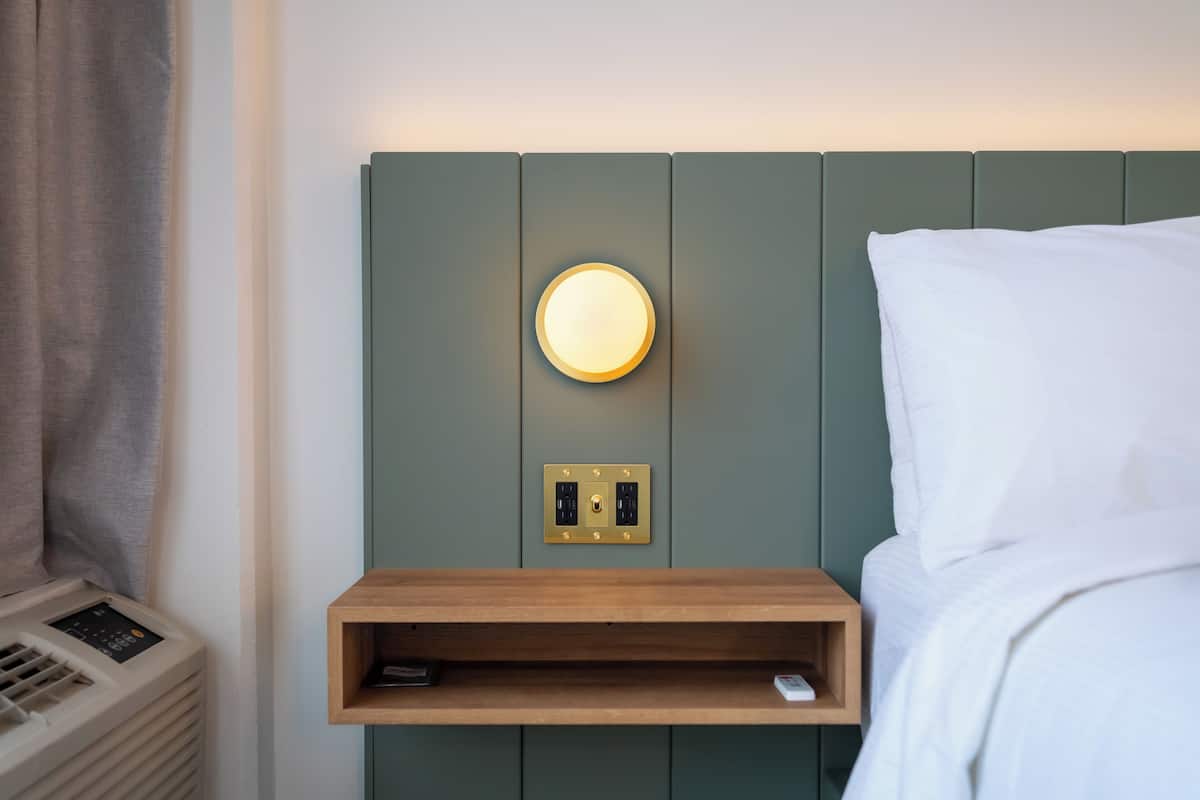 Un lit bien fait, avec une table de chevet en bois, une lampe fixée au mur et un thermostat.