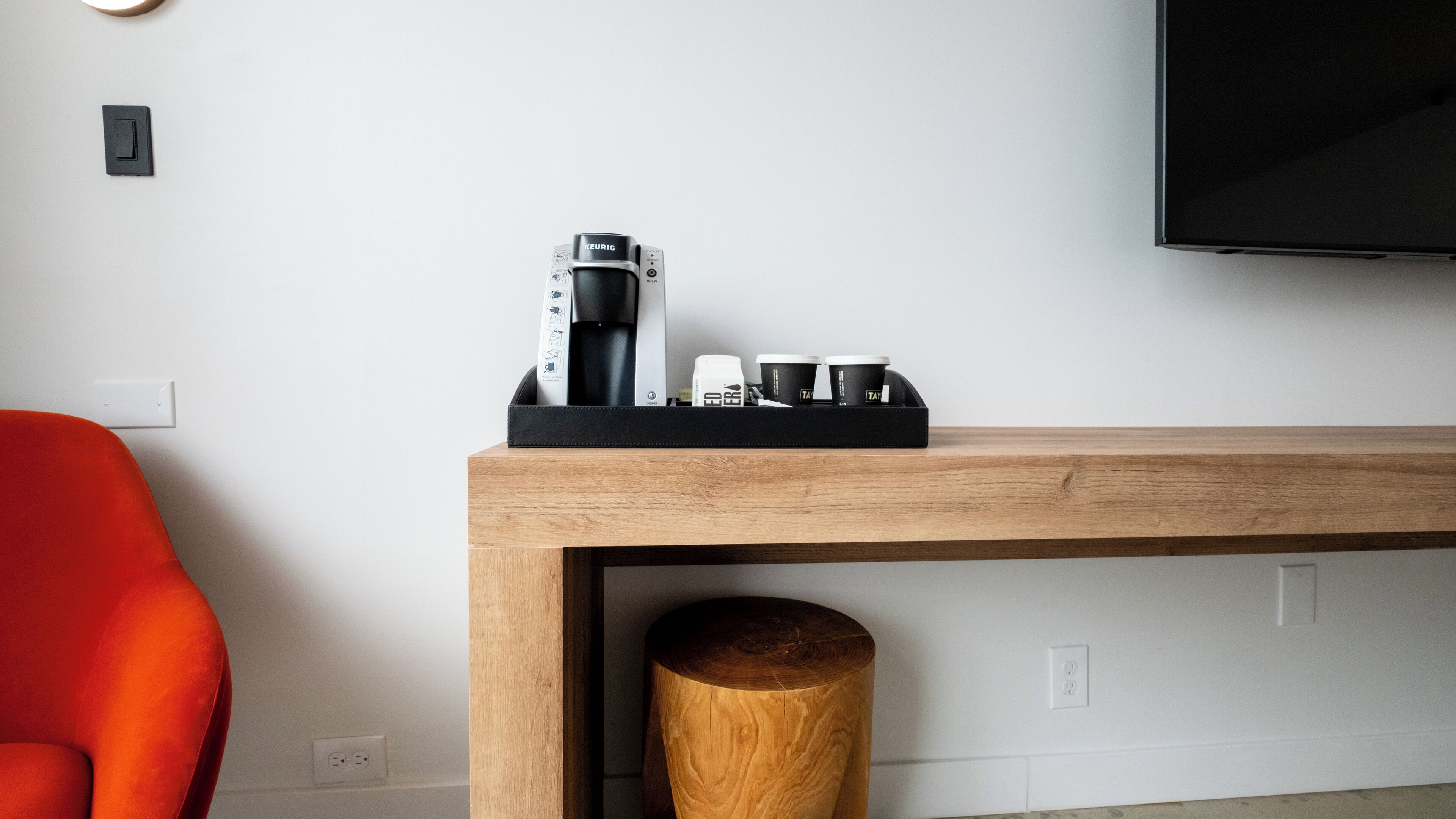 Une chambre d’hôtel moderne avec une console en bois sur laquelle sont posés une machine à café, des tasses et un plateau. Une chaise rouge est visible sur la gauche.