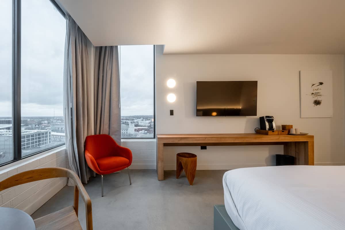 Une chambre d’hôtel avec une grande fenêtre, un bureau avec une machine à café, une chaise rouge et un lit.