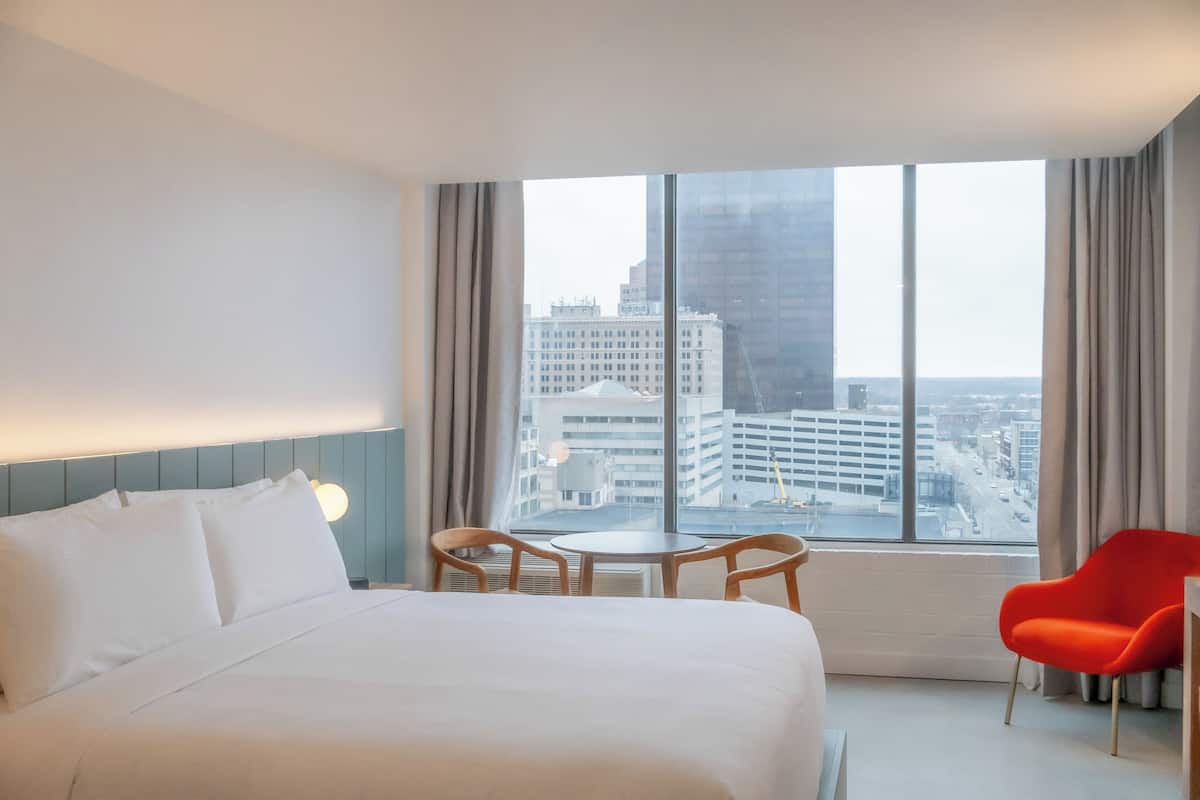 Une chambre d’hôtel avec un grand lit, une petite table, deux chaises et une vue sur les bâtiments de la ville, visible à travers de grandes fenêtres.