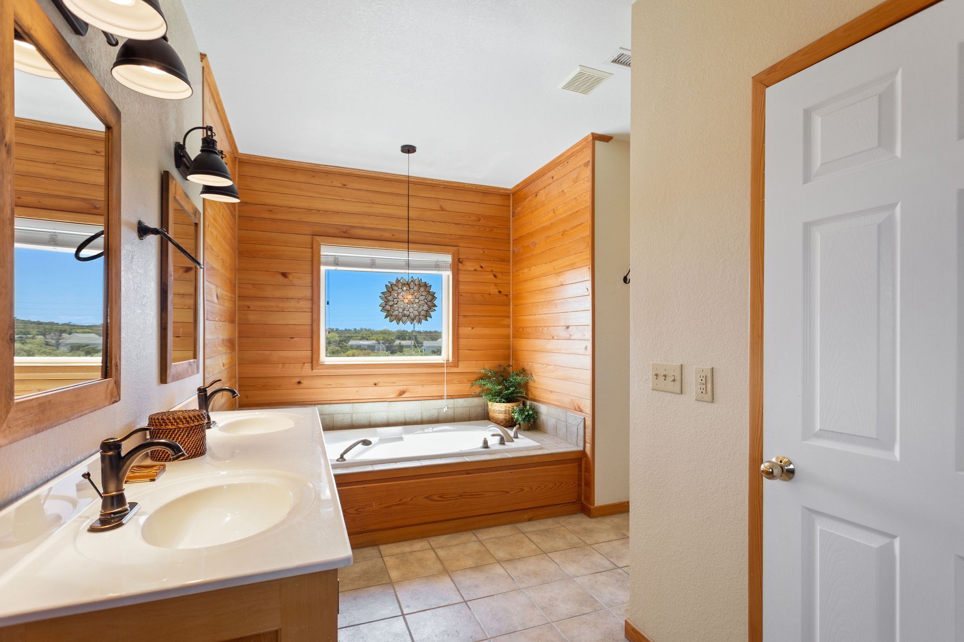 A bathroom with wooden paneling, a large mirror, two sinks, a bathtub, and a window with a decorative view.