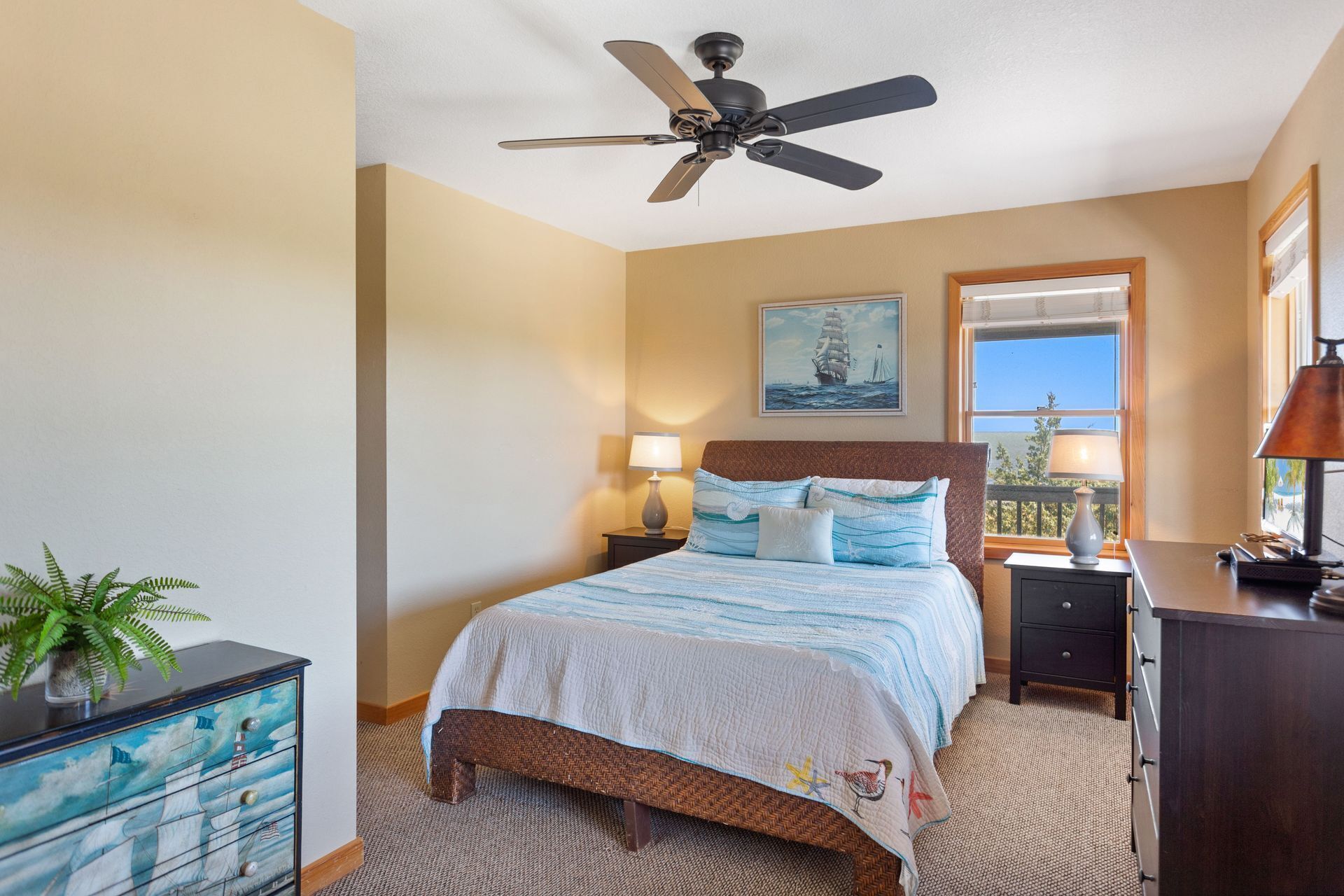 A bedroom with a bed, a ceiling fan, a nightstand with a lamp, a dresser, a window with a view, and a painting on the wall.