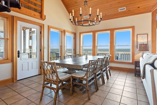 A dining area with a large table and chairs, large windows with ocean views, wooden ceiling, and a chandelier.