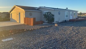 A single-story mobile home with a garage, a small deck, and a gravel driveway. - #1 Greenside Oasis (Mattawa)