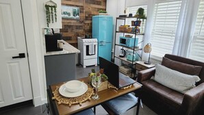 Une cuisine moderne avec un four encastré, un réfrigérateur bleu et un micro-ondes posé sur une étagère noire. On y trouve une table à manger avec un set de table et un verre de vin. Un canapé en cuir marron se trouve dans l’espace salon, agrémenté d’un coussin blanc.