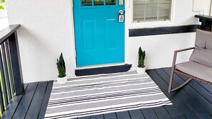 Une porte turquoise avec une pancarte indiquant « bonjour », une fenêtre, un banc noir et un tapis à rayures sur une terrasse en bois.