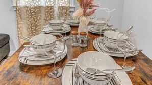 Una mesa de comedor con platos, tazones y cubiertos blancos, junto con copas de vino y un florero decorativo con flores.