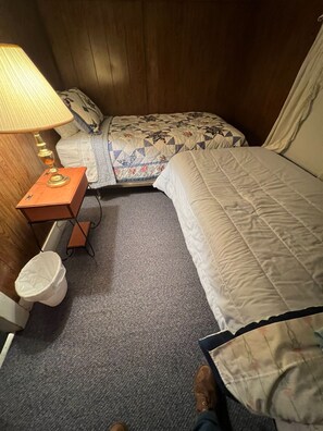 A bedroom with two beds, a wooden headboard, a nightstand with a lamp, and a trash bin.