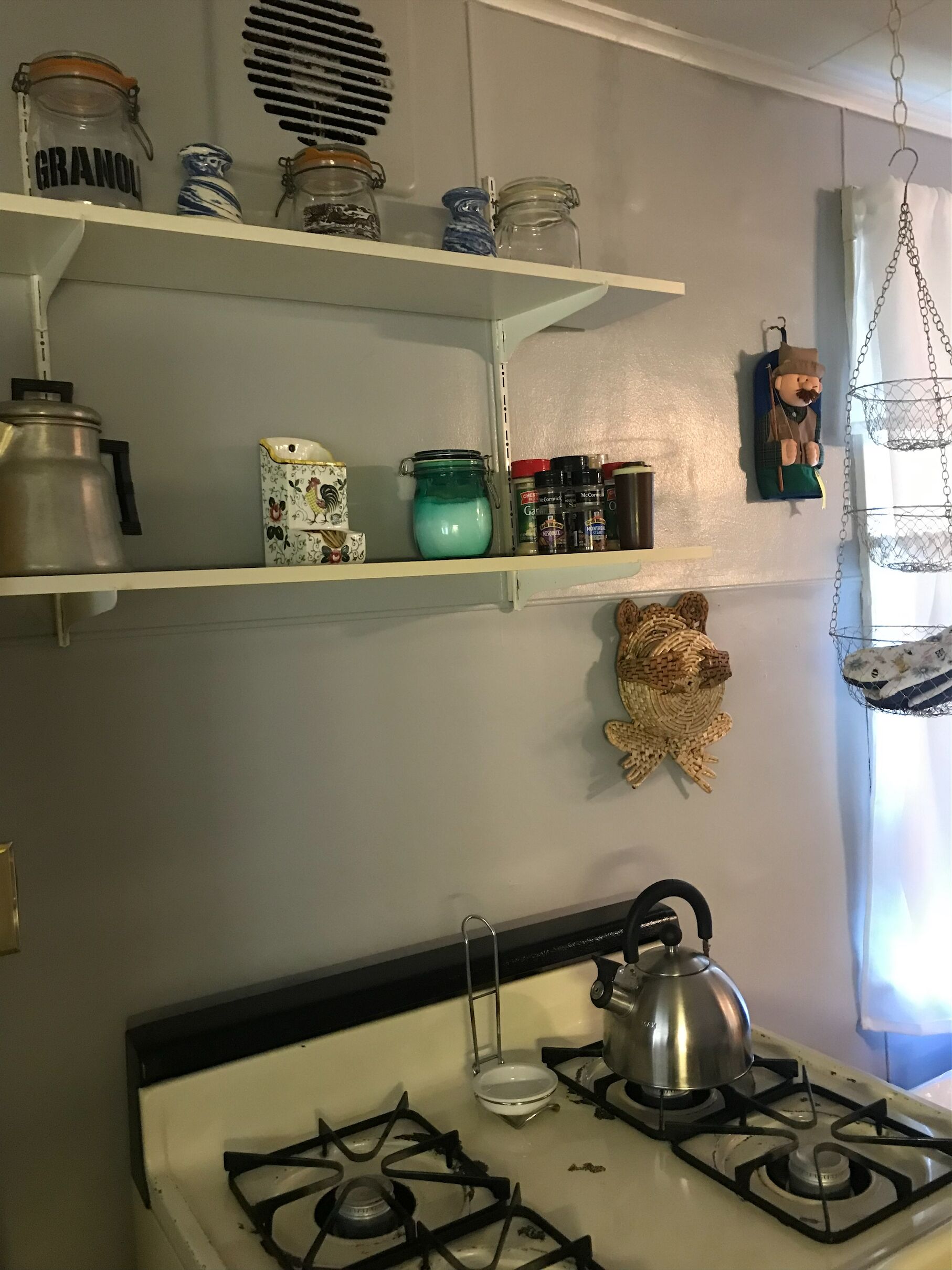 A kitchen with a gas stove, shelves with jars, and a hanging decorative item.