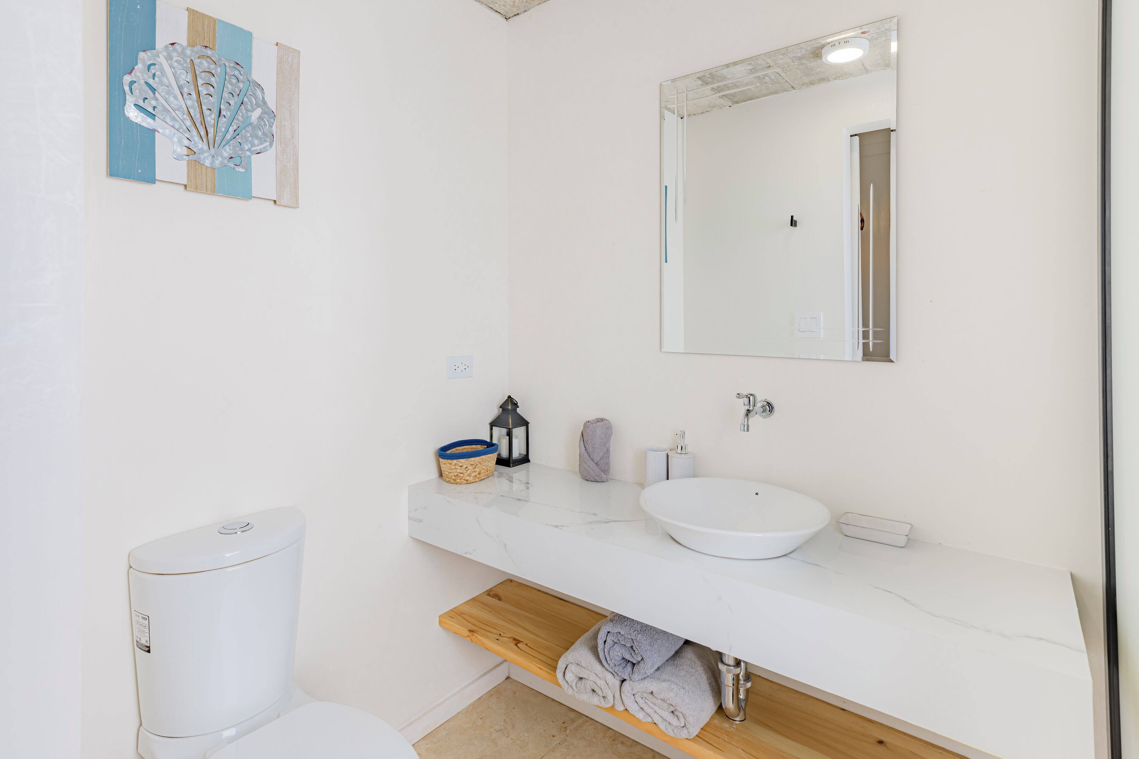 A modern bathroom with a white toilet, a sink with a mirror above, and a wooden shelf with towels.