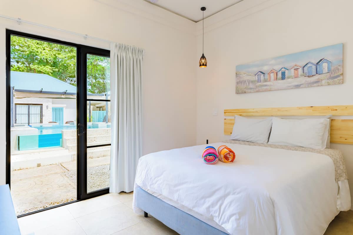 Een slaapkamer met een groot bed, een schilderij van strandhuisjes en uitzicht op een zwembad door een glazen deur.