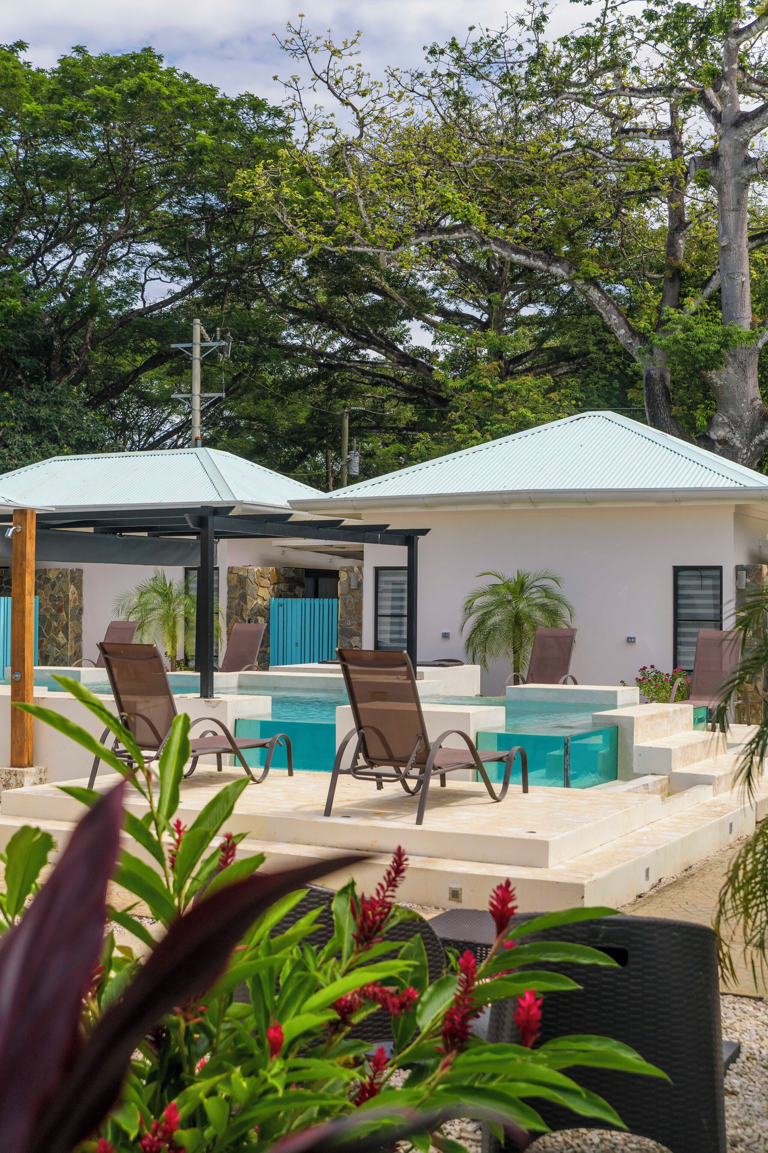 A pool area with lounge chairs, a covered seating area, and a building with a light-colored roof.