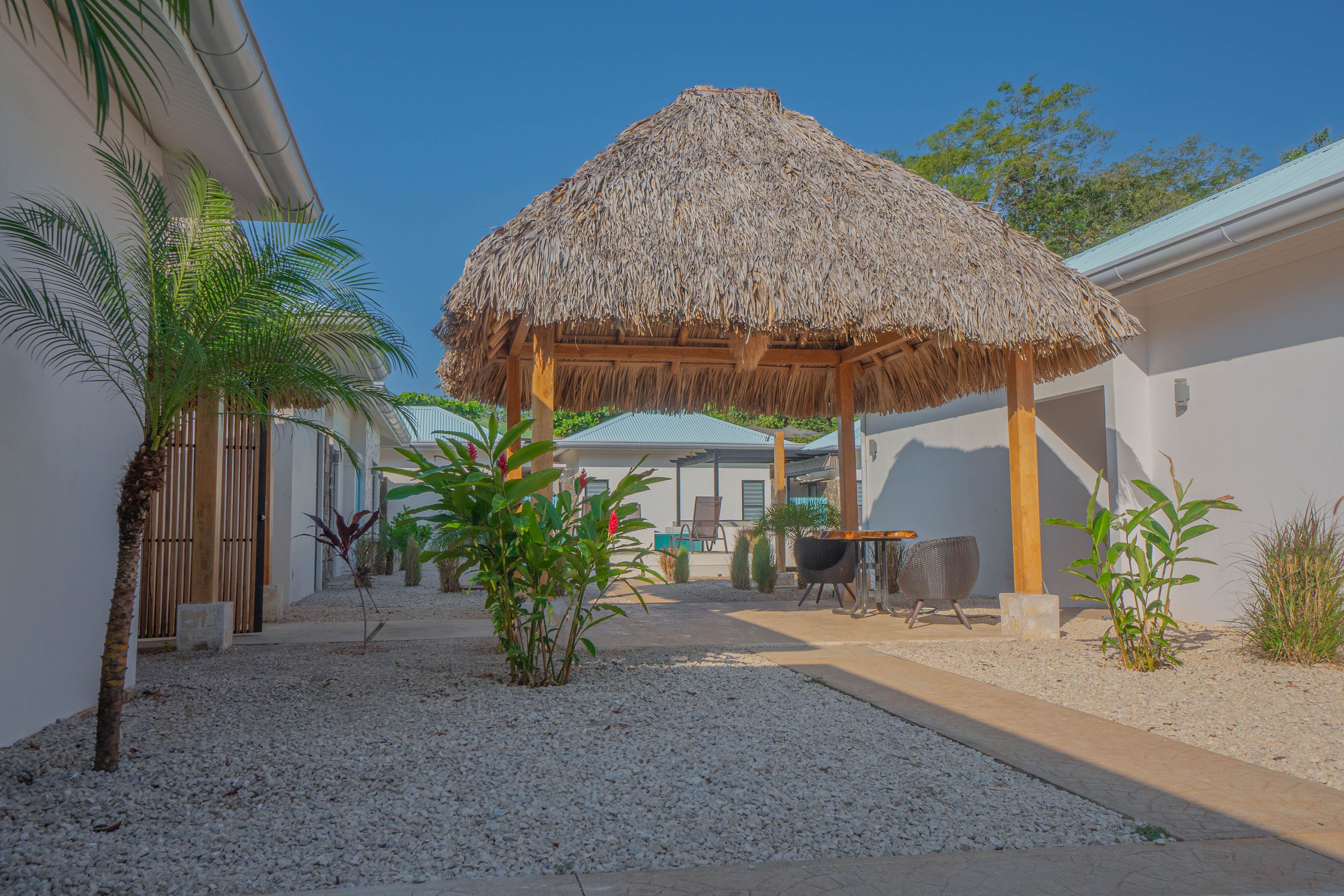 A covered outdoor seating area with a thatched roof, surrounded by palm trees and plants, and a gravel path leading to a building.