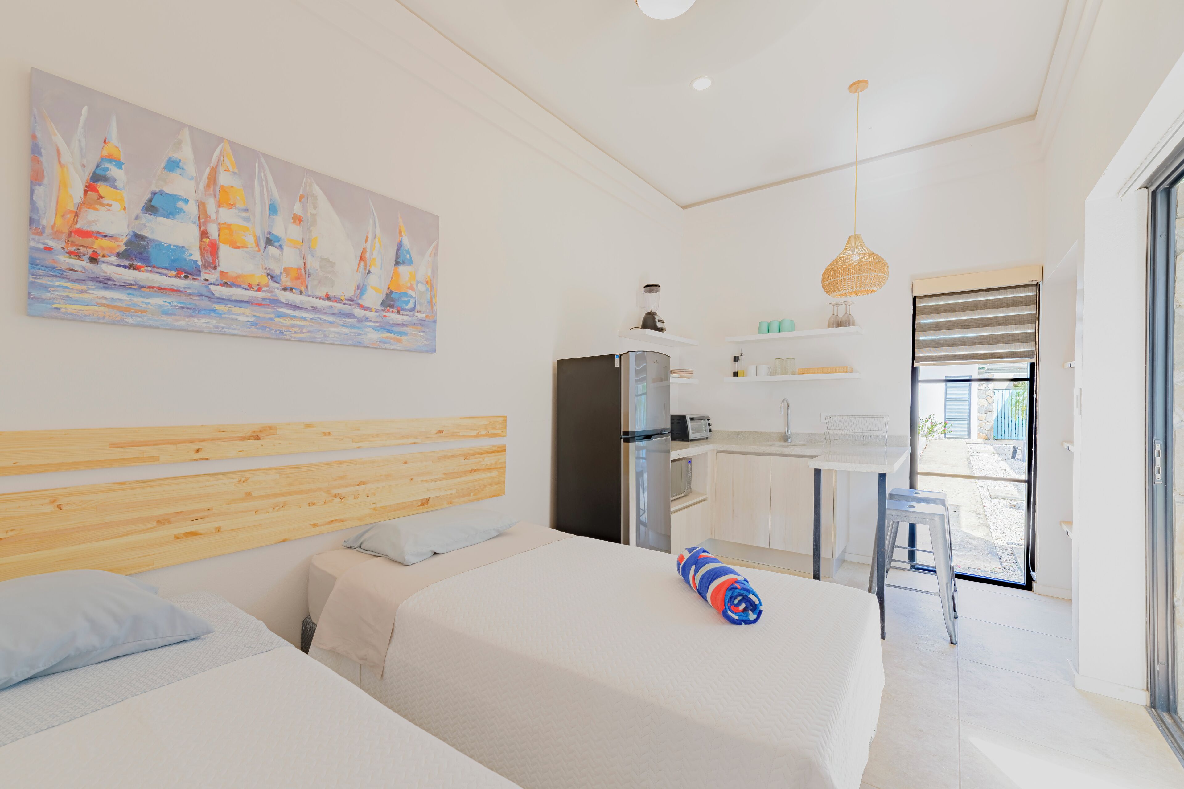 Ein modernes Hotelzimmer mit einem Holzkopfteil, einem Bett mit weißer Bettdecke, einem Segelbootgemälde an der Wand, einer Küchenzeile und einer Schiebetür zum Außenbereich.