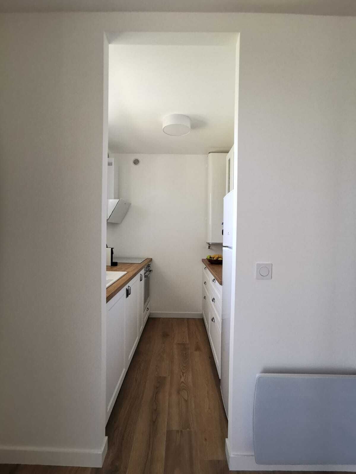A narrow, well-lit hallway with a wooden floor, leading to a kitchen area with white cabinets and a wooden countertop.