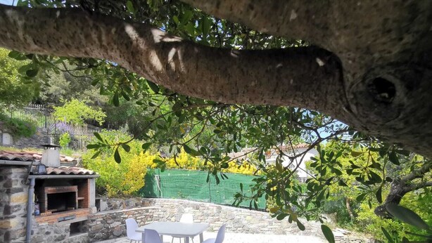A stone patio with a table and chairs, a brick oven, and a green fence in the background. - Bas de Villa au Calme (Villeneuve-Loubet)
