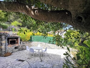 A stone patio with a table and chairs, a brick oven, and a green fence in the background. - Bas de Villa au Calme (Villeneuve-Loubet)